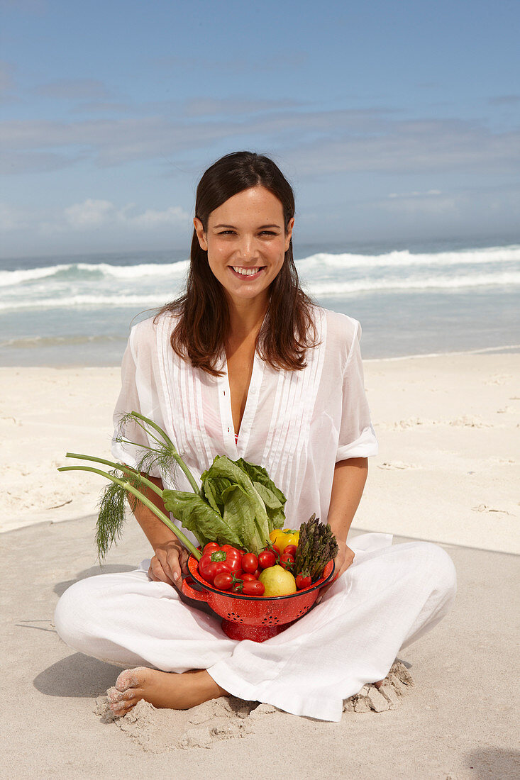 A young brunette woman on a beach wearing a white summer dress and holding a bowl of vegetables