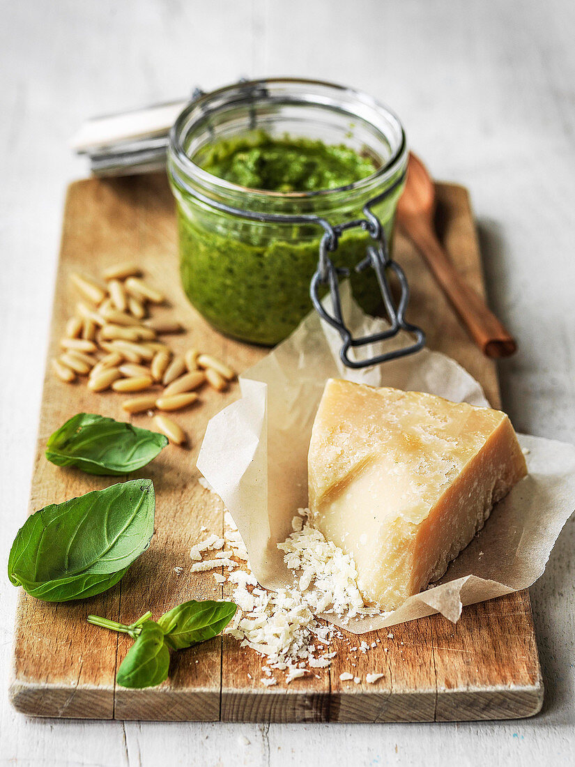 A jar of pesto with parmesan cheese pine nuts and basil on board