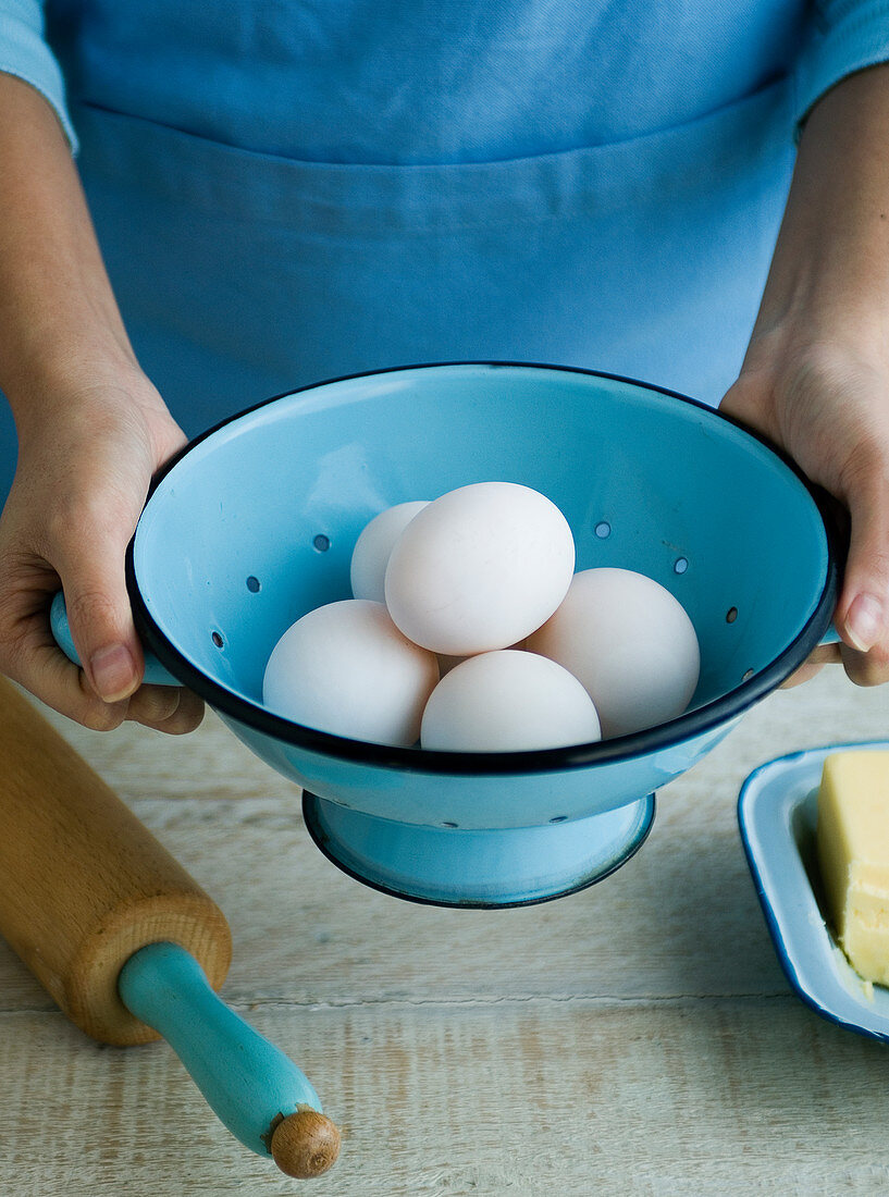 Person hält blauen Emailleseiher mit weißen Eiern daneben Teigroller und Butter