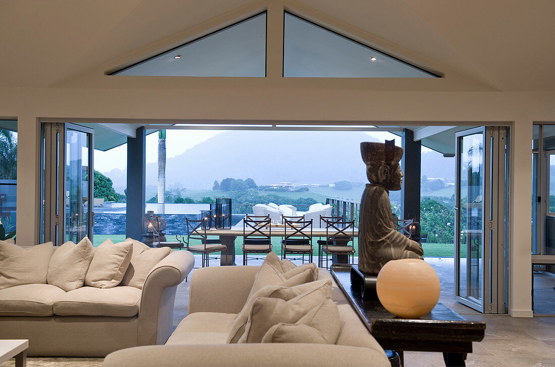 Elegante Polstergarnitur und Konsole mit Buddha-Figur in Lounge, im Hintergrund Terrasse mit Landschaftsblick