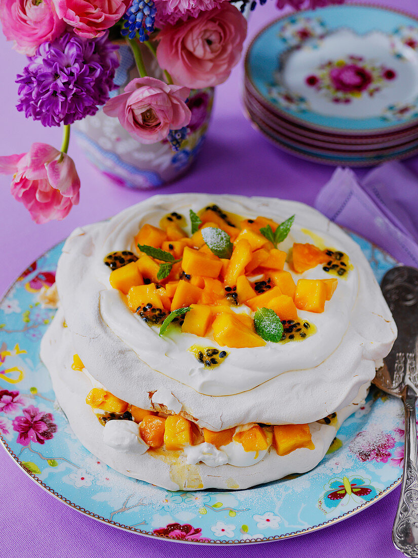 Pavlova mit Mango und Passionsfrucht auf geblümtem Teller, im Hintergrund Blumenstrauß