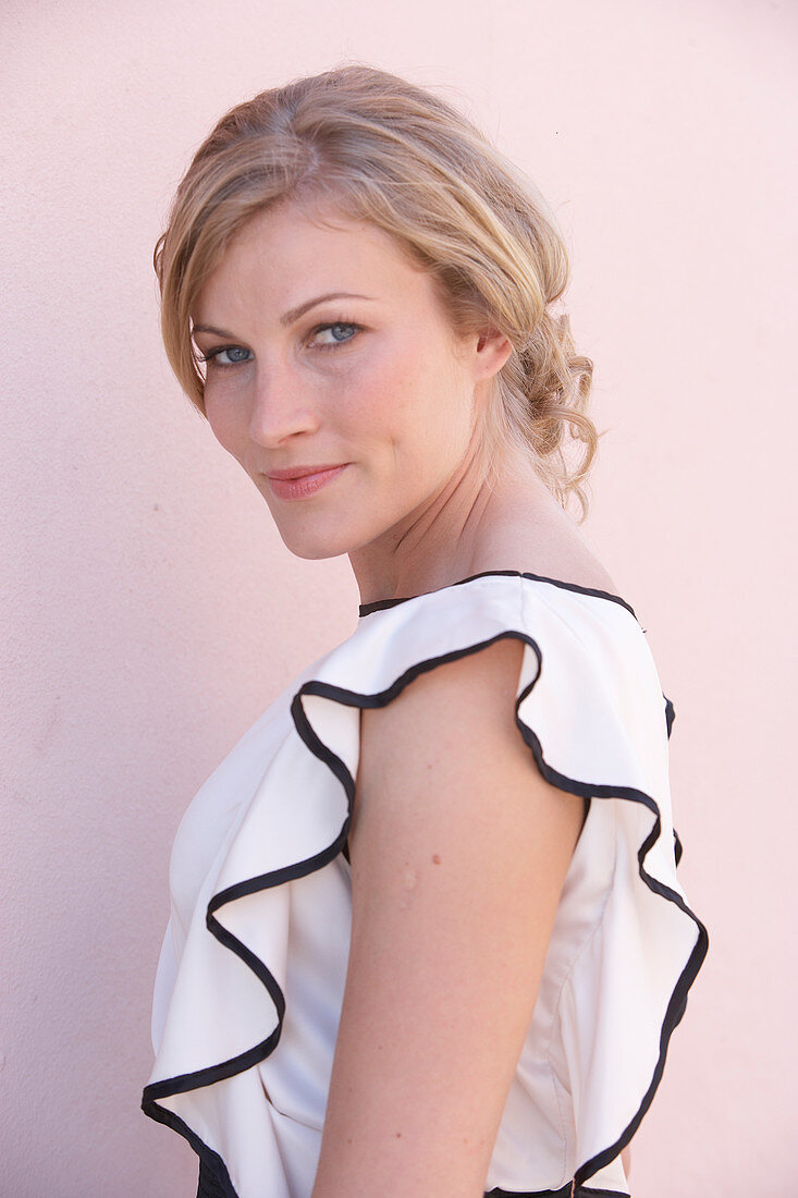 A blonde woman standing against a pink background wearing a light blouse with a black trim