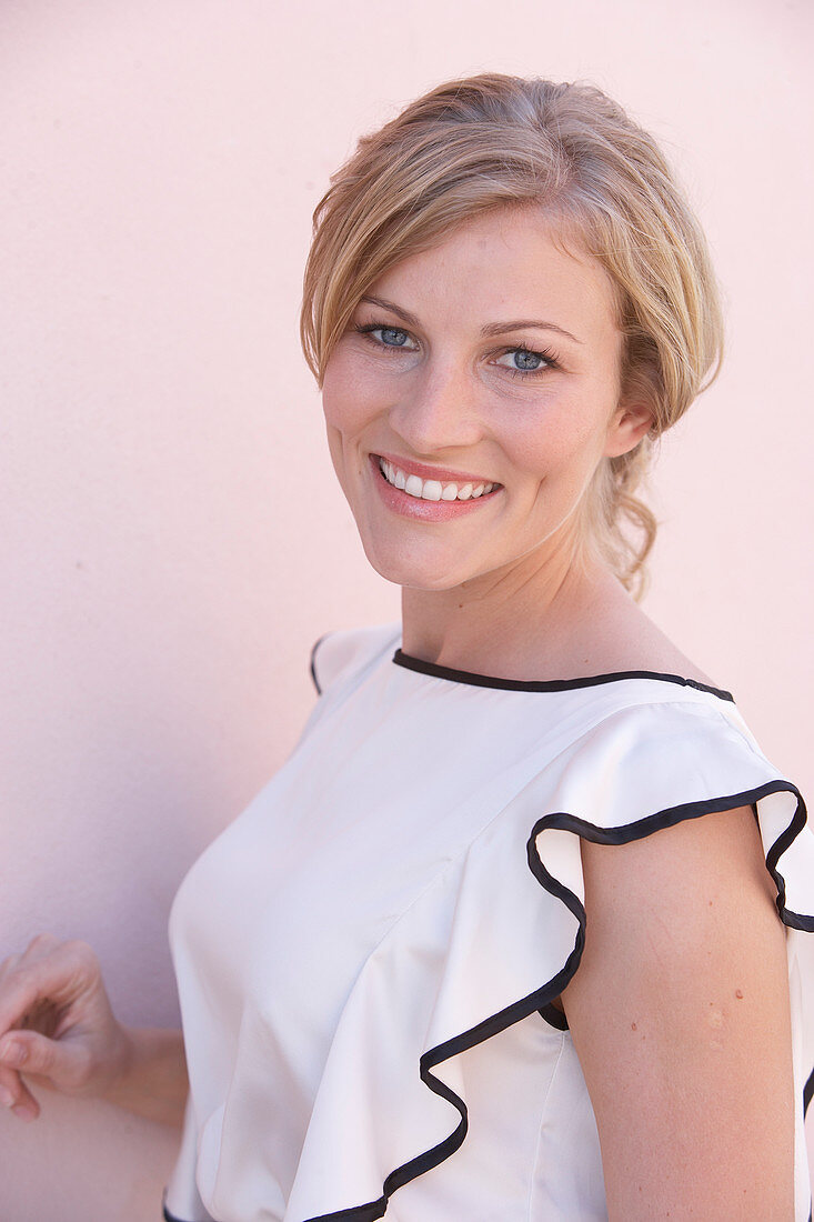 A blonde woman standing against a pink background wearing a light blouse with a black trim