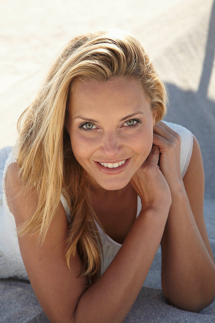 A mature blonde woman lying on a sandy beach wearing a white top