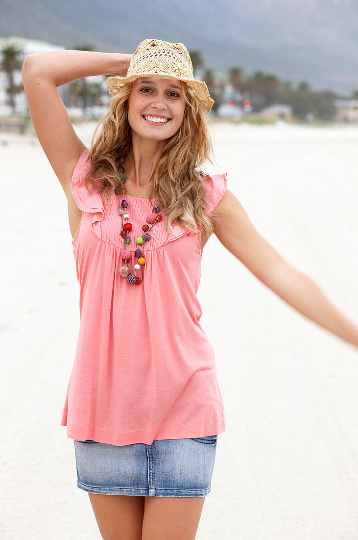 A young blonde woman on a beach wearing a pink top, a short denim skirt and a beige hat