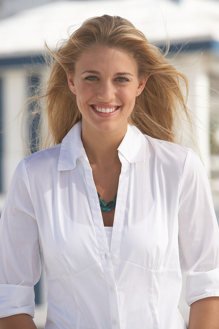 A young blonde woman on a beach wearing a white blouse and a chunky necklace