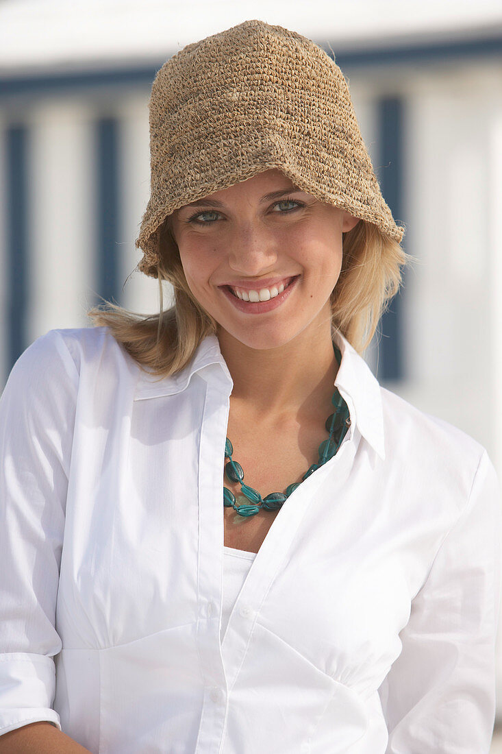 A young blonde woman on a beach wearing a white blouse and a brown hat