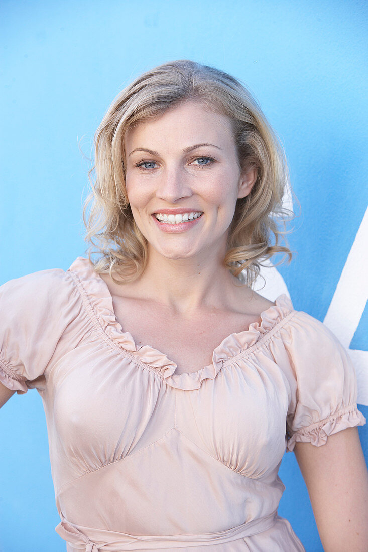 A blonde woman standing against a blue surface wearing a pink short-sleeved blouse