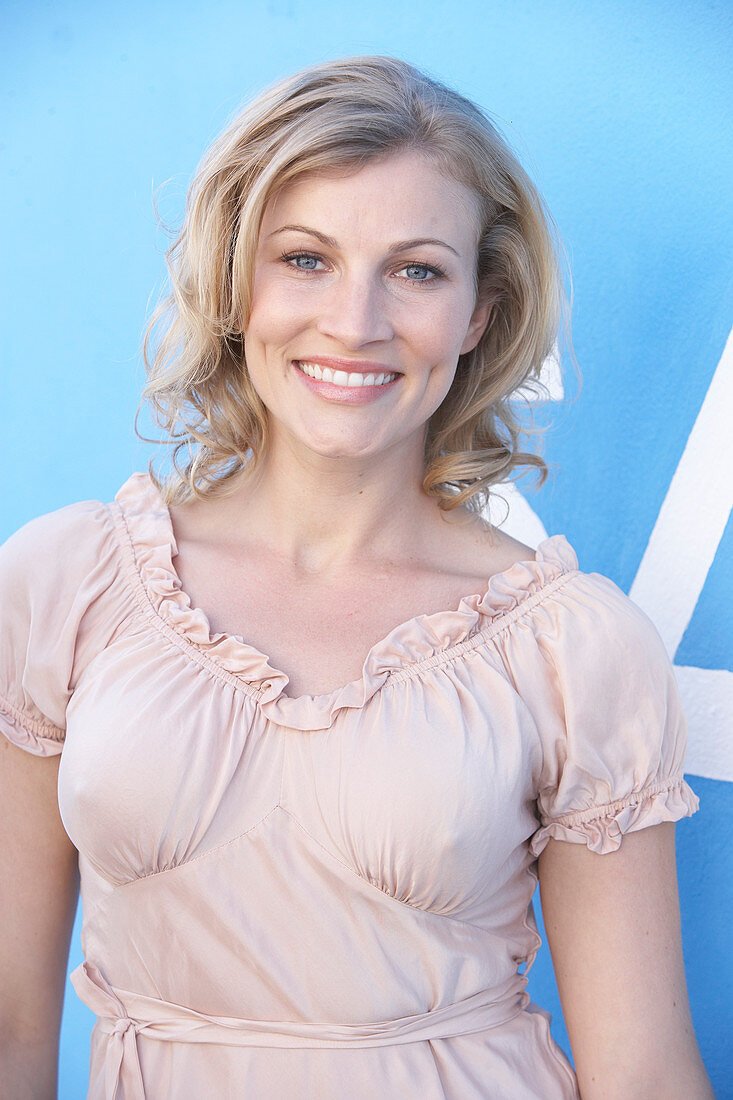 A blonde woman standing against a blue surface wearing a pink short-sleeved blouse