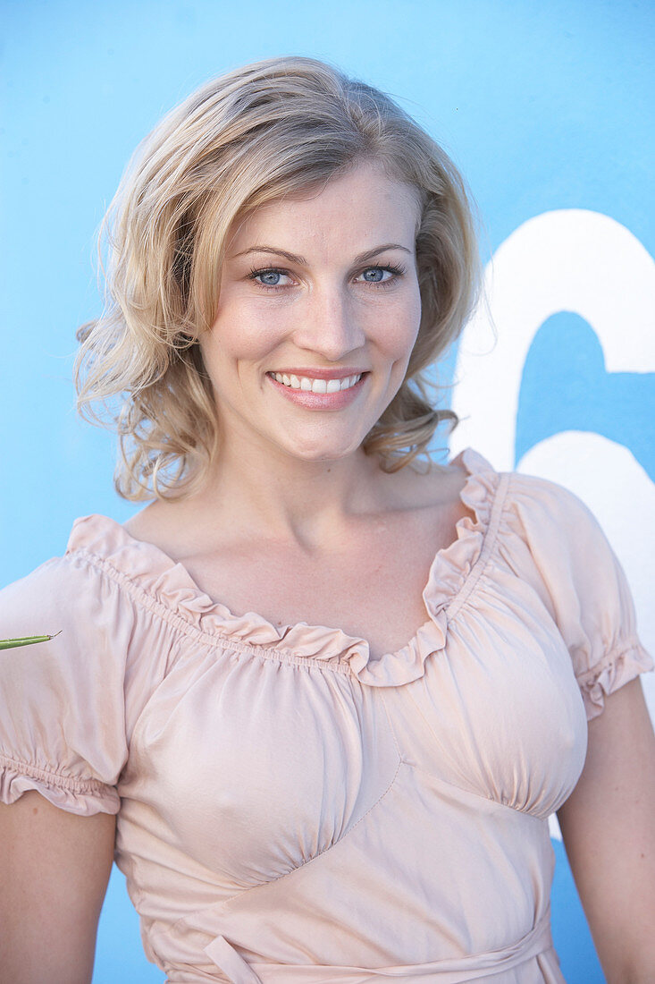 A blonde woman standing against a blue surface wearing a pink short-sleeved blouse