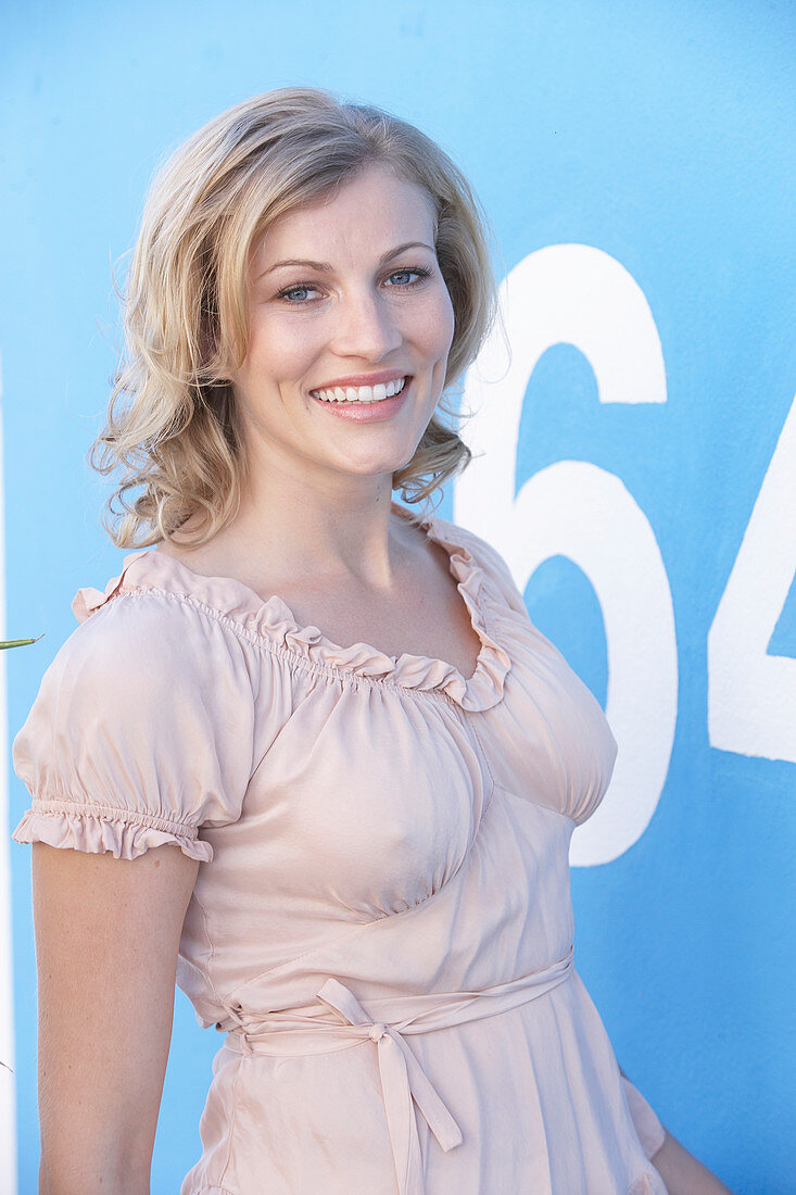 A blonde woman standing against a blue surface wearing a pink short-sleeved blouse