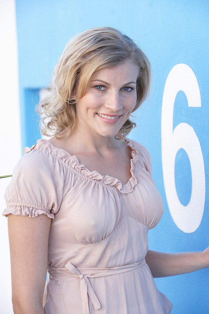 A blonde woman standing against a blue surface wearing a pink short-sleeved blouse