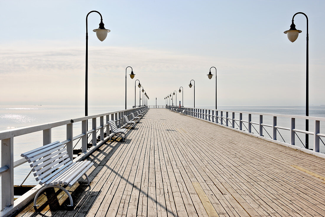 Ocean pier with balustrade, street lamps and benches below blue sky
