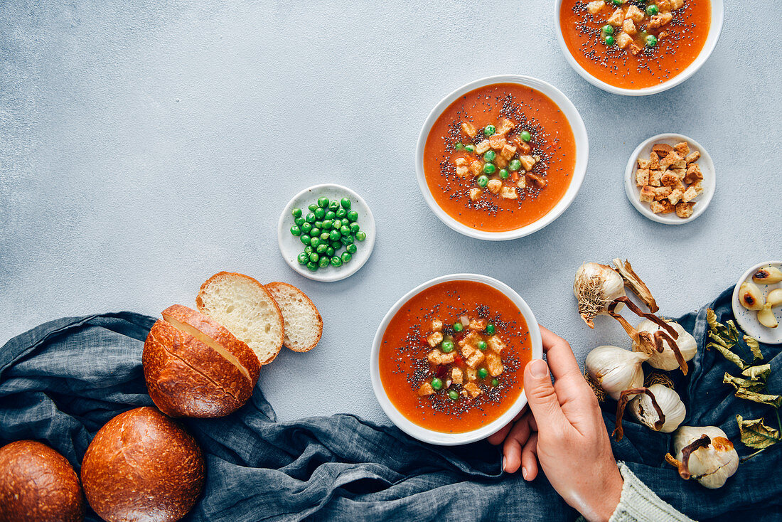 Vegan tomato soup served in three white bowls accompanied by bread rolls, roasted garlic, croutons and peas
