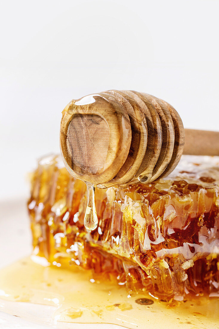 Organic honey in honeycombs flowing from wooden dipper on white marble background