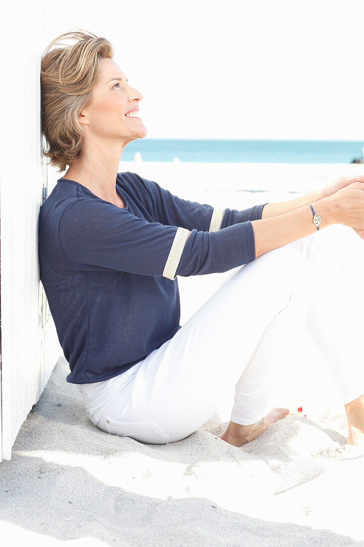 A mature woman with short blonde hair on a beach wearing a blue shirt and white trousers
