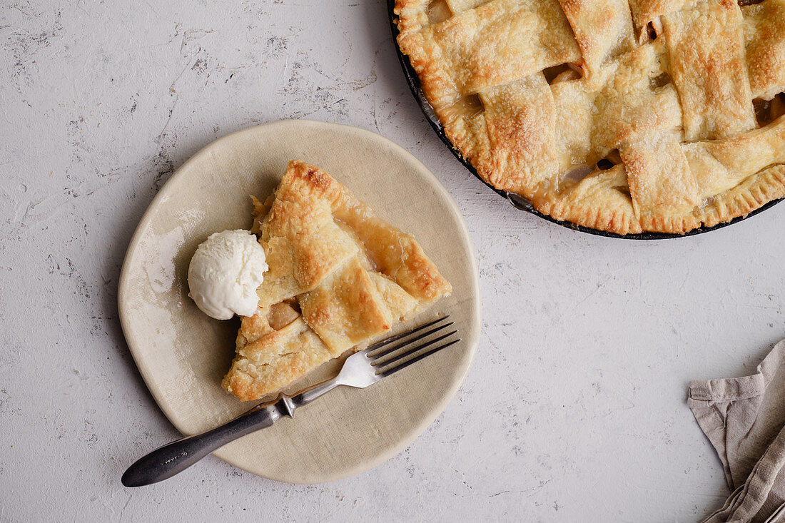 Apple pie with lattice decoration and a slice served on handmade ceramic plate with a scoop of vanilla ice-cream