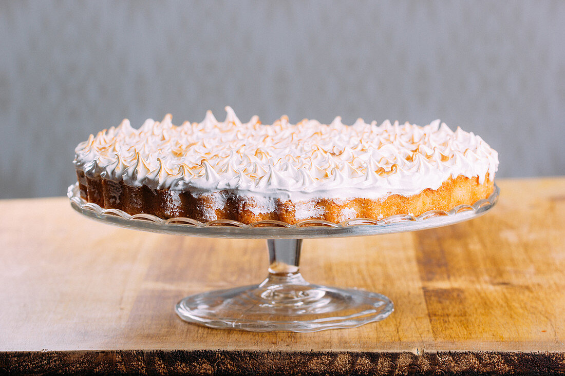 Crispy tart topped with baked white fluffy meringue and served on glass cake stand on table