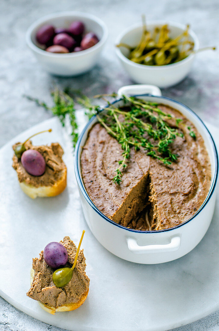 Liver pate in ceramic form with fresh thyme, and two baguette sandwiches with pate, capers and olives on a marble stand