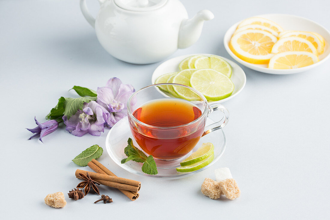 Tee in Glastasse daneben Zucker, Gewürze, Blüten und Fruchtscheiben