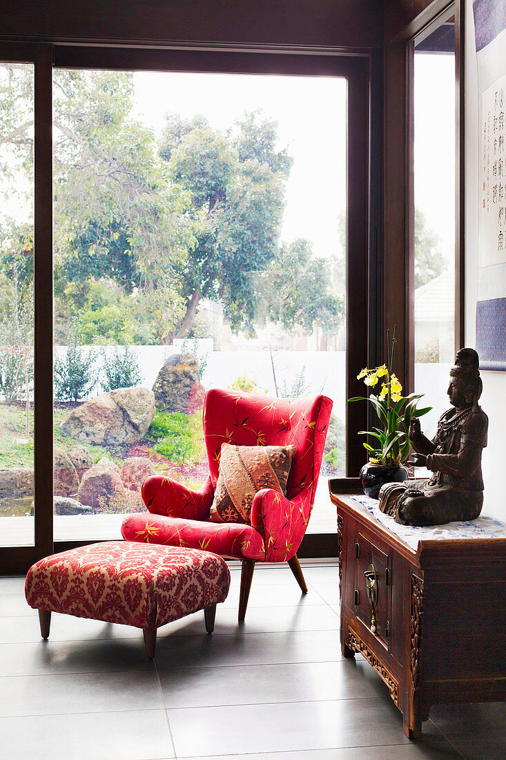 Red wing chair in fifties style, with matching footstool, antique cupboard with Buddha figure in front of floor-to-ceiling glass door