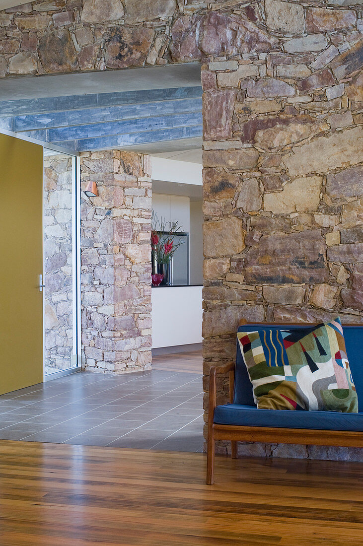 Wooden sofa with blue upholstery and colourful scatter cushion next to open doorway in stone house