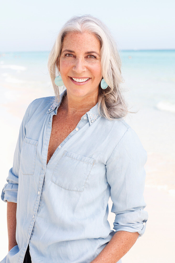 A mature woman with white hair on a beach wearing a light blue blouse