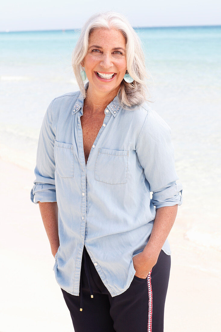Reife Frau mit weißen Haaren in hellblauer Bluse und dunkelblauer Hose am Strand