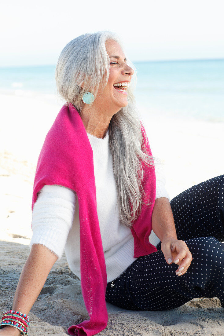 A mature woman with white hair on a beach wearing a white jumper and polka dot trousers with a pink jumper over her shoulders