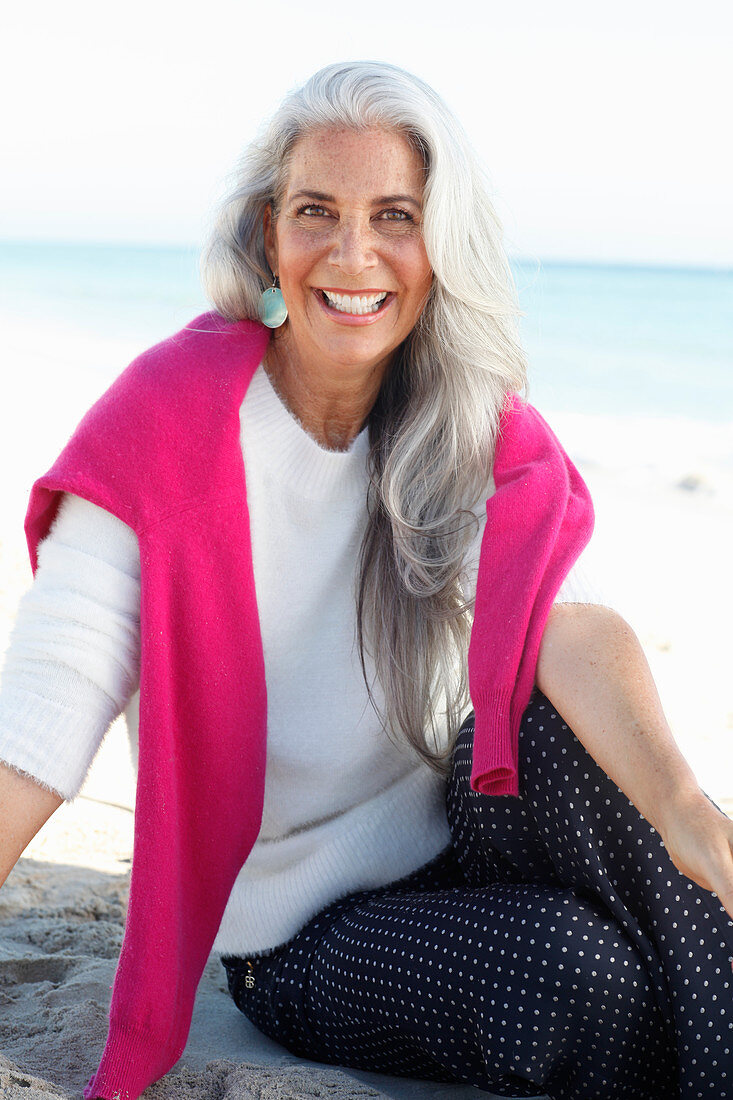 A mature woman with white hair on a beach wearing a white jumper and polka dot trousers with a pink jumper over her shoulders