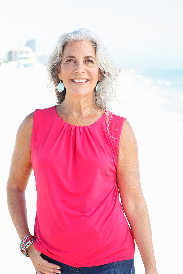 A mature woman with white hair on a beach wearing a pink top and blue jeans