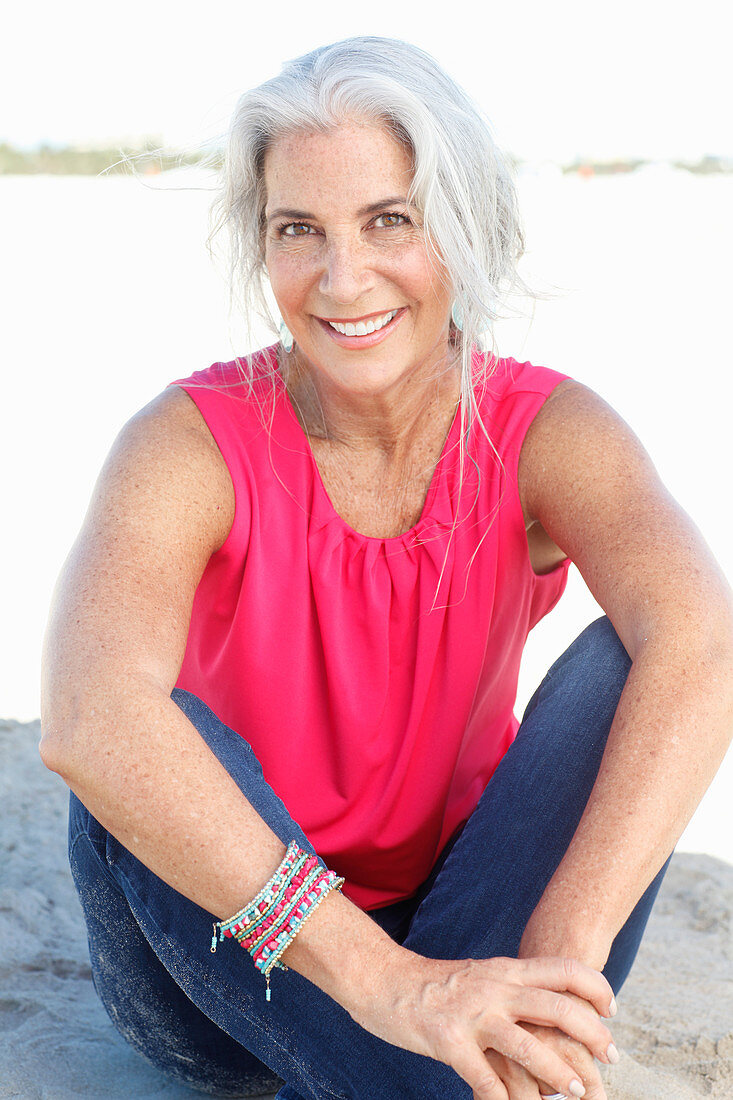 A mature woman with white hair on a beach wearing a pink top and blue jeans