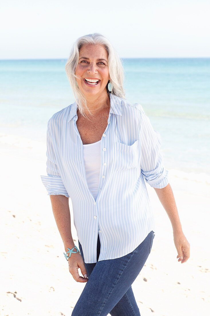 Reife Frau mit weißen Haaren in gestreifter Bluse, Top und Bluejeans am Strand