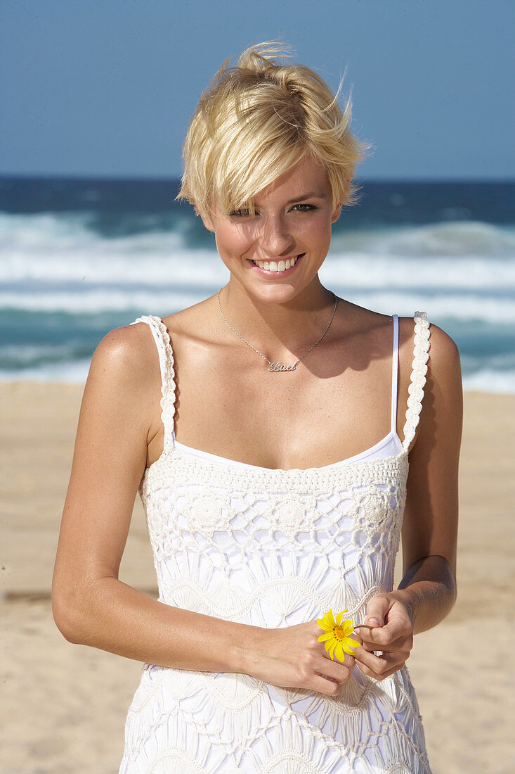 A blonde woman with short hair on a beach wearing a white dress