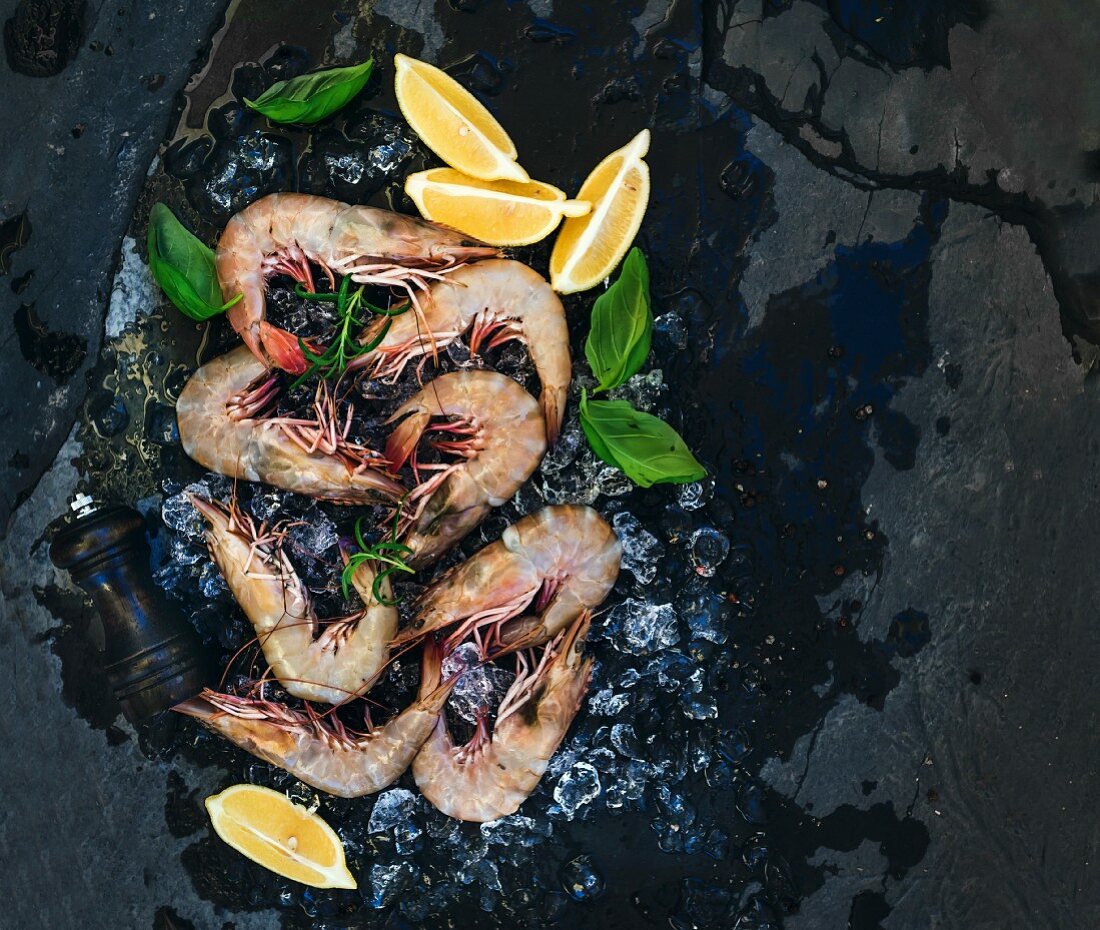 Fresh uncooked shrimps with lemon, herbs and spices on chipped ice over dark slate stone backdrop