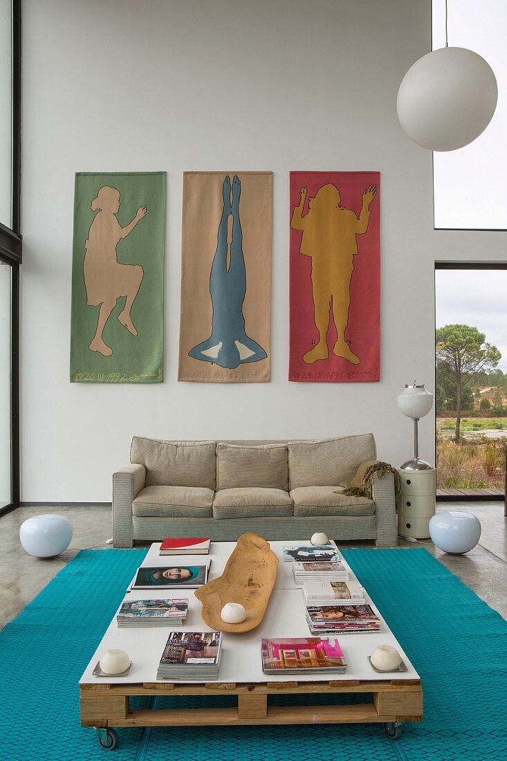 Pallet table on castors and sofa below three wall hangings in high-ceilinged interior