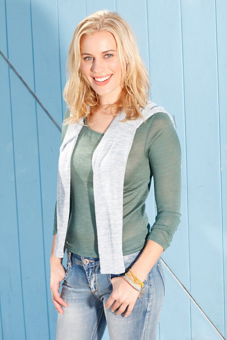 Blonde Frau in grünem Strick-Shirt, grauem Pullover über den Schultern und Jeans