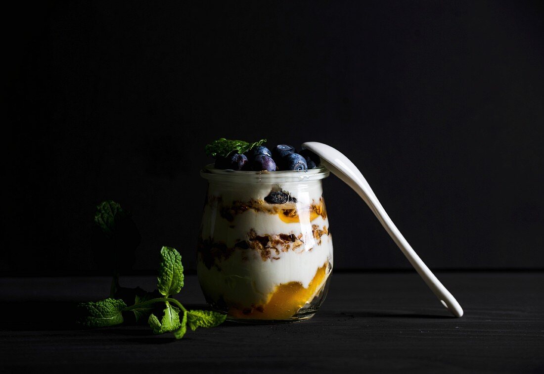 Hafermüsli mit Joghurt, Marmelade, Heidelbeeren und Minze im Glas vor schwarzer Hintergrund