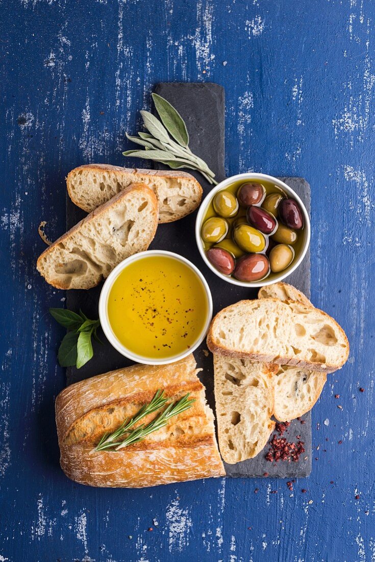 Mediterranean snacks set: Olives, oil, herbs and sliced ciabatta bread on black slate stone board