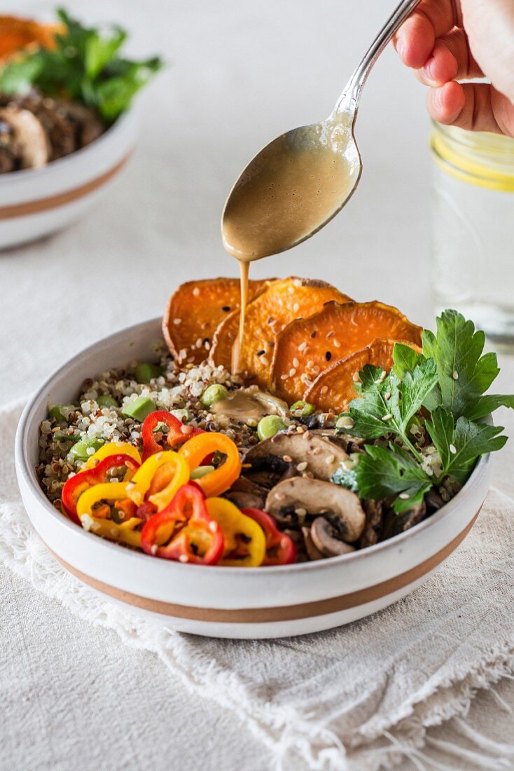 A bowl of quinoa topped with sweet potatoes, mushrooms, bell peppers, and drizzled with a tahini sauce by a person