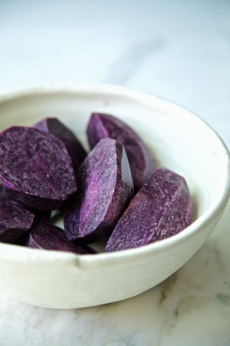 Purple potatoes, cut and peeled in a bowl, on a white marble background