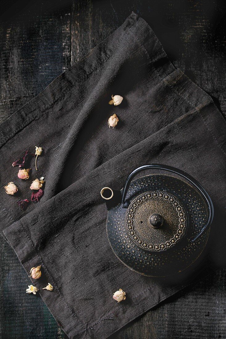 Schwarze, asiatische Teekanne aus Gusseisen und getrocknete Rosenknospen auf schwarzem Textil