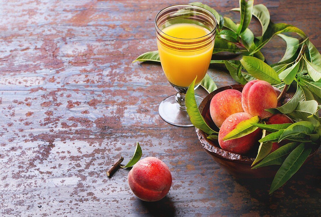 Peaches on branch with leaves in wooden bowl and glass with peach s juice over old metal background