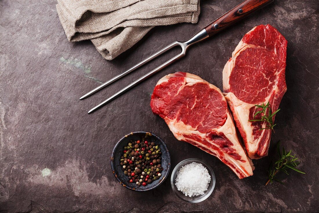Zwei rohe Ribeye-Steaks, herzförmig arrangiert, mit Rosmarin, Pfeffer und Salz auf Schieferplatte