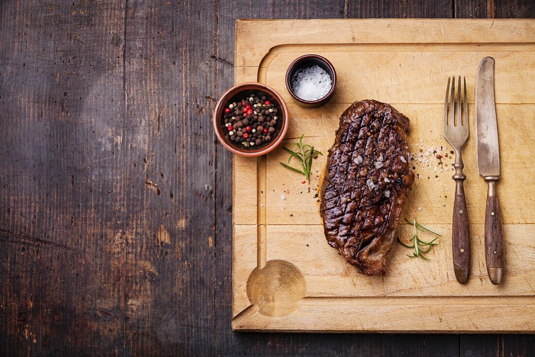Grilled New York Striploin Steak with salt and pepper on meat cutting board on dark wooden background