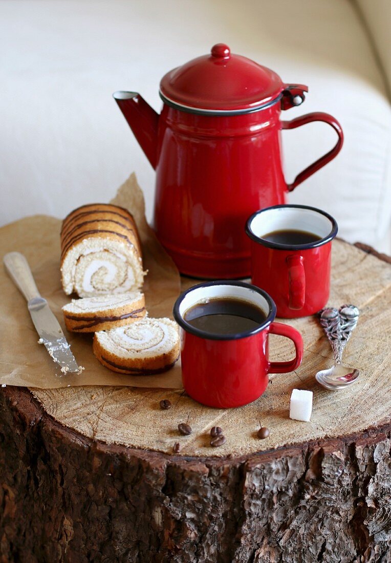 Coffee in red enamel cups and a jug served with a Swiss roll cake on a tree trunk