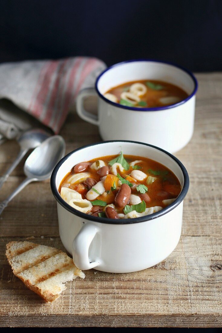 Two enamel mugs of minestrone soup with pasta, beans and vegetables