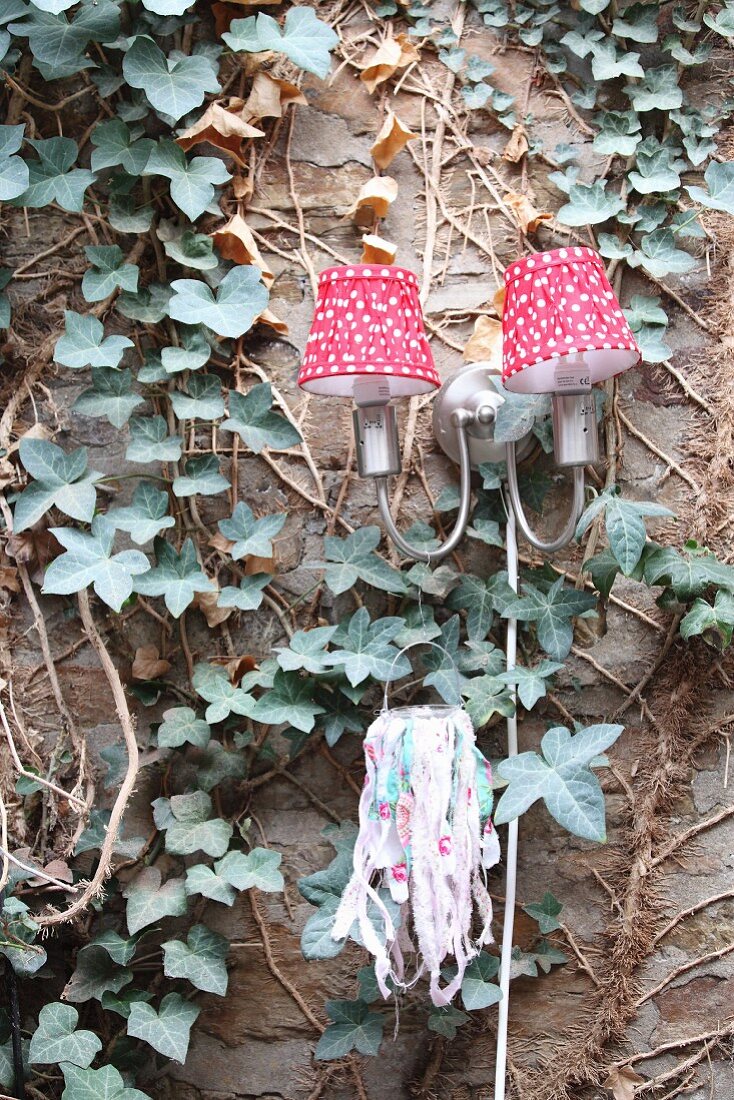 Sconce lamp with red and white lampshades and tealight holder on ivy-covered wall