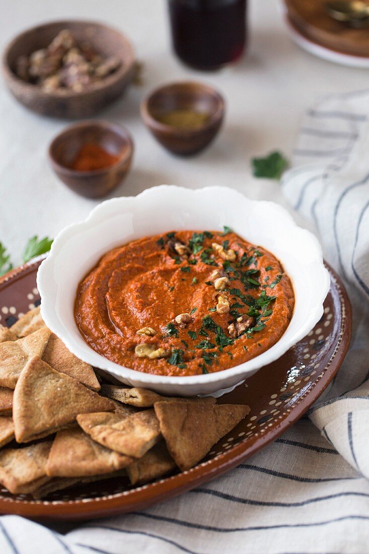 A bowl of red pepper and walnut dip served with pita chips on a plate