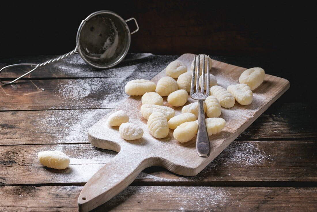Uncooked homemade potato gnocchi with fork and strainer on vintage cutting board over wooden table with flour